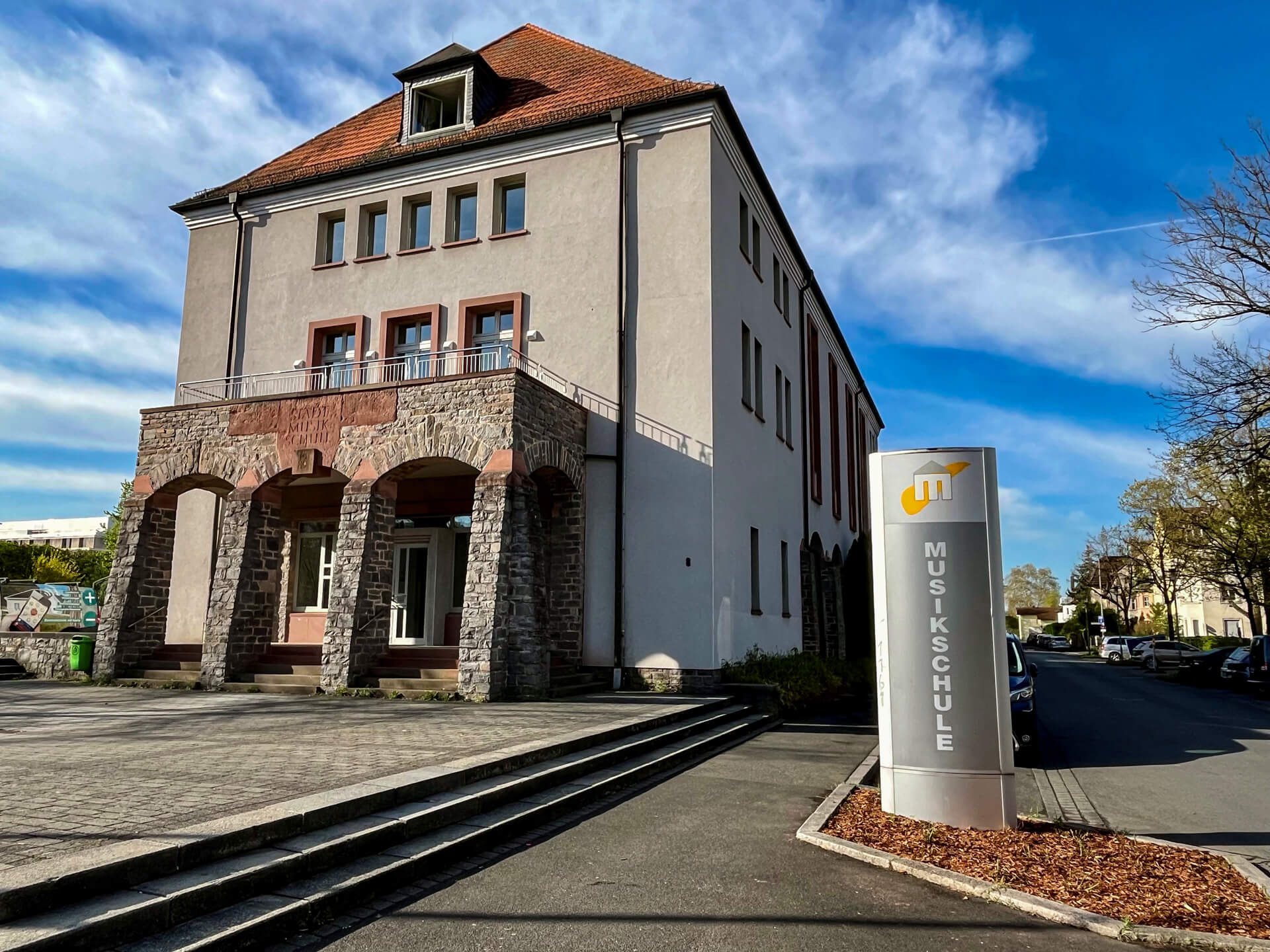 Die Außenansicht der Städtischen Musikschule Aschaffenburg an einem sonnigen Tag. Das mehrstöckige, historische Gebäude hat einen markanten Eingangsbereich mit Arkaden aus rotem Sandstein. Im Vordergrund rechts steht eine moderne Stele mit dem Logo und der senkrechten Aufschrift "MUSIKSCHULE".