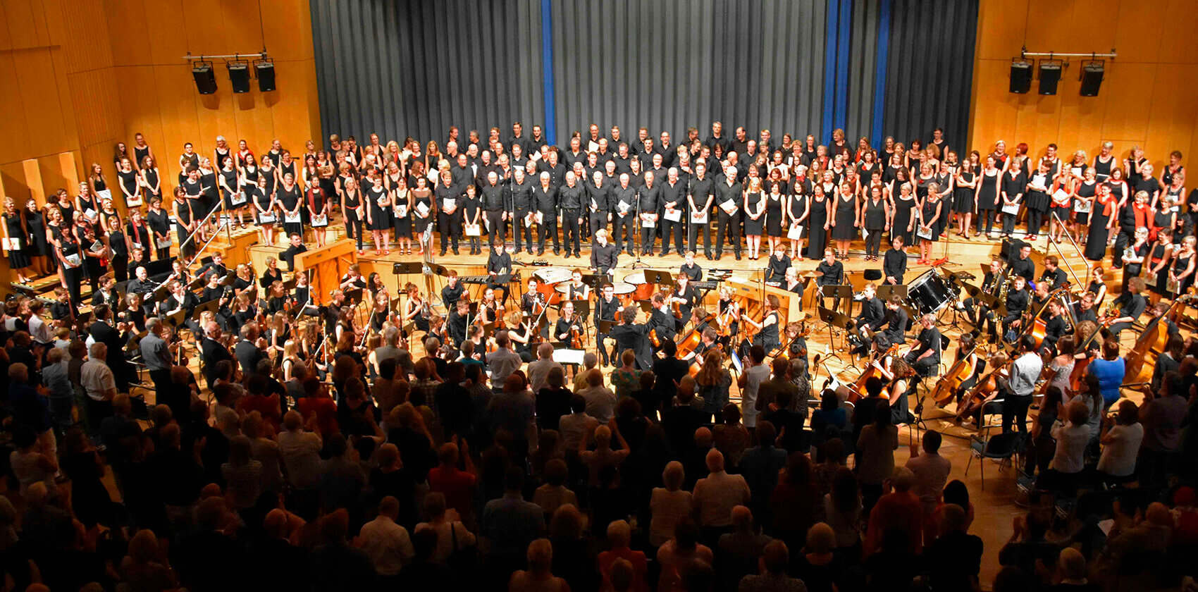 Ein Blick von oben in einen voll besetzten Konzertsaal. Auf der Bühne musizieren ein großes Orchester und ein dahinter aufgestellter, riesiger Chor gemeinsam. Im Vordergrund ist das Publikum zu sehen, das der Aufführung zusieht.