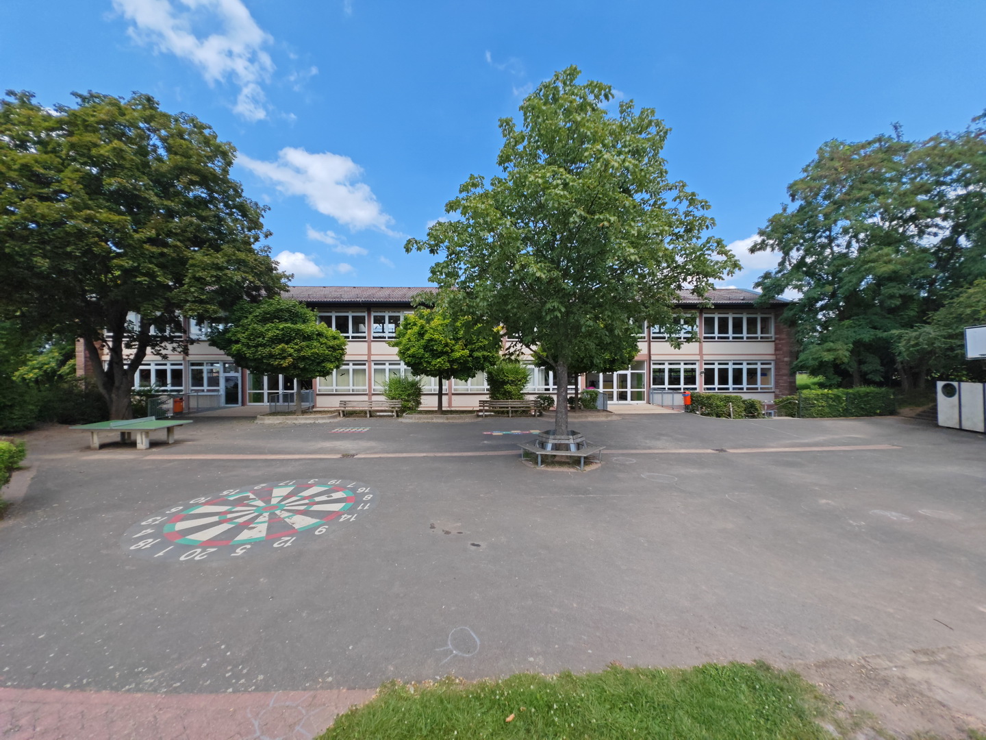 Der sonnige Schulhof der Astrid-Lindgren-Grundschule. Auf dem asphaltierten Boden ist ein großes, buntes Spiel in Form einer Zielscheibe aufgemalt. Mehrere große Laubbäume spenden Schatten, einer davon ist von einer Rundbank umgeben. Im Hintergrund steht das zweistöckige Schulgebäude unter einem blau-weißen Himmel.