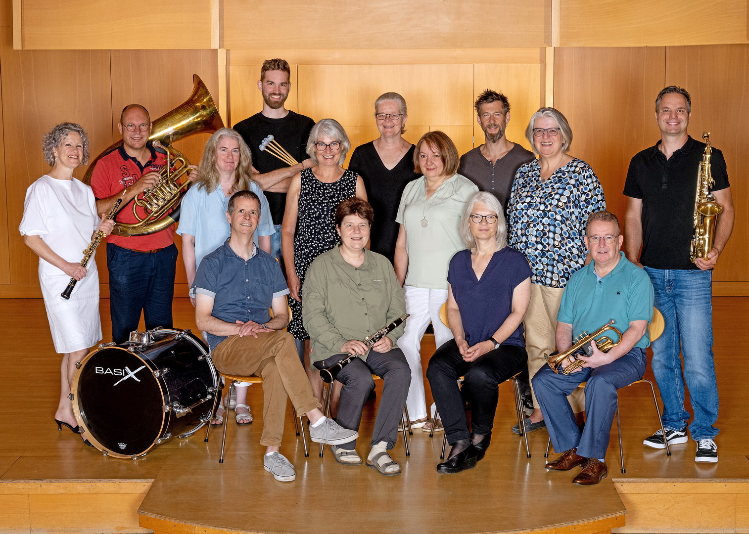 Ein Gruppenfoto der Lehrkräfte für Blasinstrumente. Eine Gruppe von Männern und Frauen ist auf einer kleinen Bühne mit ihren Instrumenten aufgestellt, darunter eine Tuba, ein Saxofon, eine Klarinette und eine Trompete.