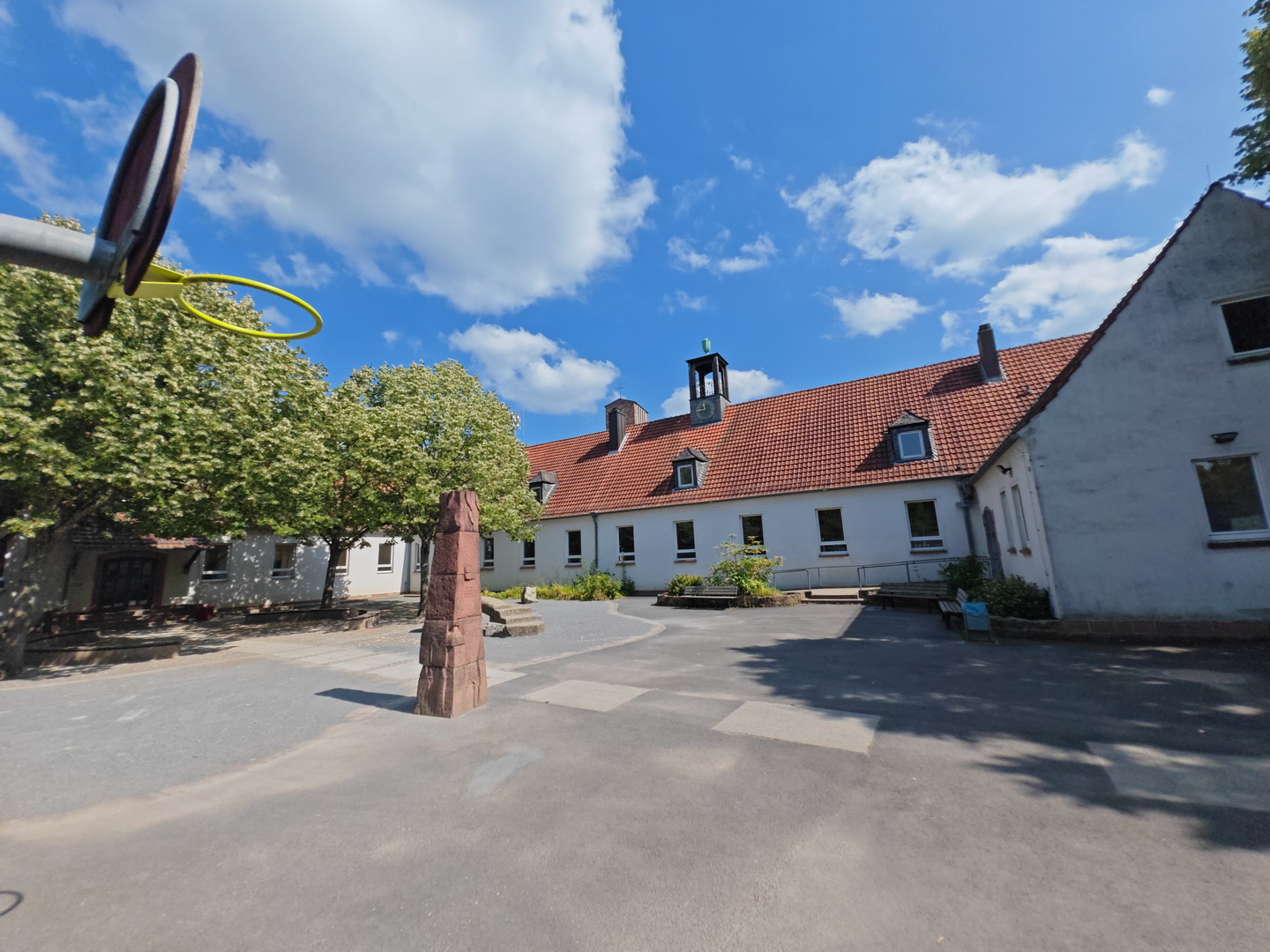 Ein sonniger Blick in den Innenhof der Christian-Schad-Schule. Der Hof ist von weißen Gebäuden mit einem durchgehenden roten Ziegeldach umgeben, auf dem in der Mitte ein kleiner Glockenturm sitzt. Im Hof stehen große Laubbäume und eine rote Sandstein-Stele, während links ein Basketballkorb ins Bild ragt. Der Himmel ist blau mit vielen weißen Wolken.