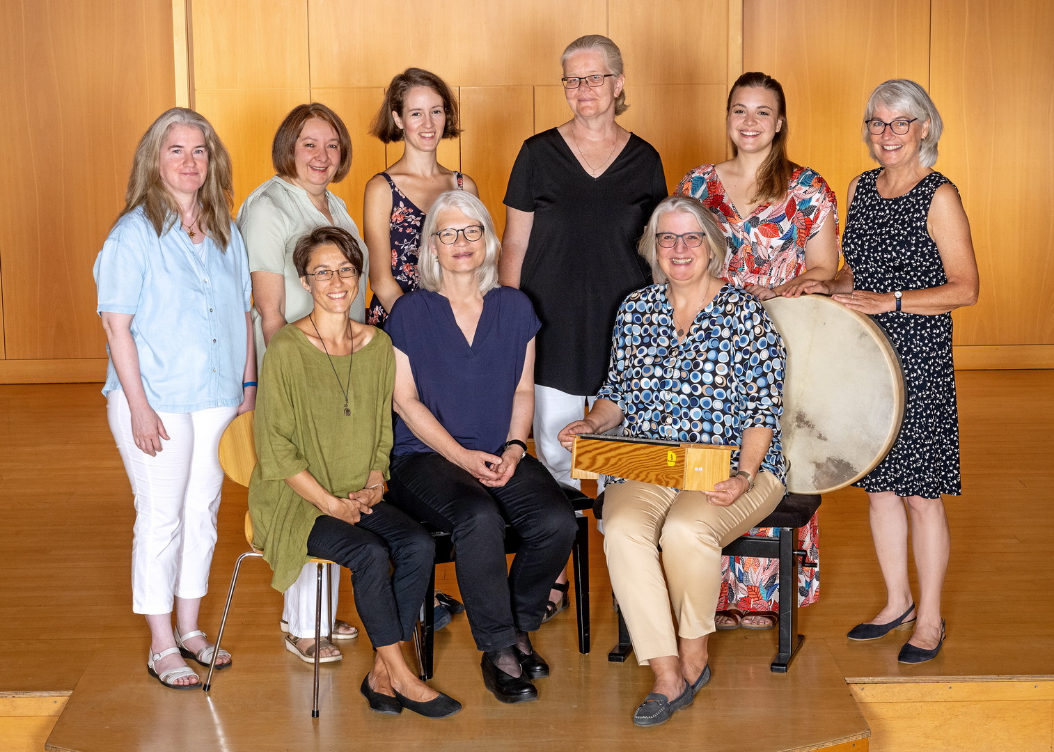 Ein Gruppenfoto der neun Lehrerinnen für Elementare Musikerziehung. Die Frauen posieren teils sitzend, teils stehend in einem holzvertäfelten Saal. Einige von ihnen halten Percussion-Instrumente wie eine große Rahmentrommel in den Händen.