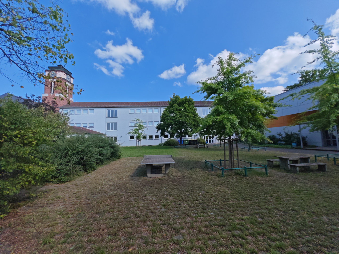 Der Blick über den grünen Schulhof der Erthalschule auf das Schulgebäude. Im Vordergrund befindet sich eine Rasenfläche mit jungen Bäumen und einer steinernen Tischtennisplatte. Das Hauptgebäude ist ein langer, heller Bau, an den sich auf der linken Seite ein markanter, historischer Turm aus rotem Gestein anschließt.