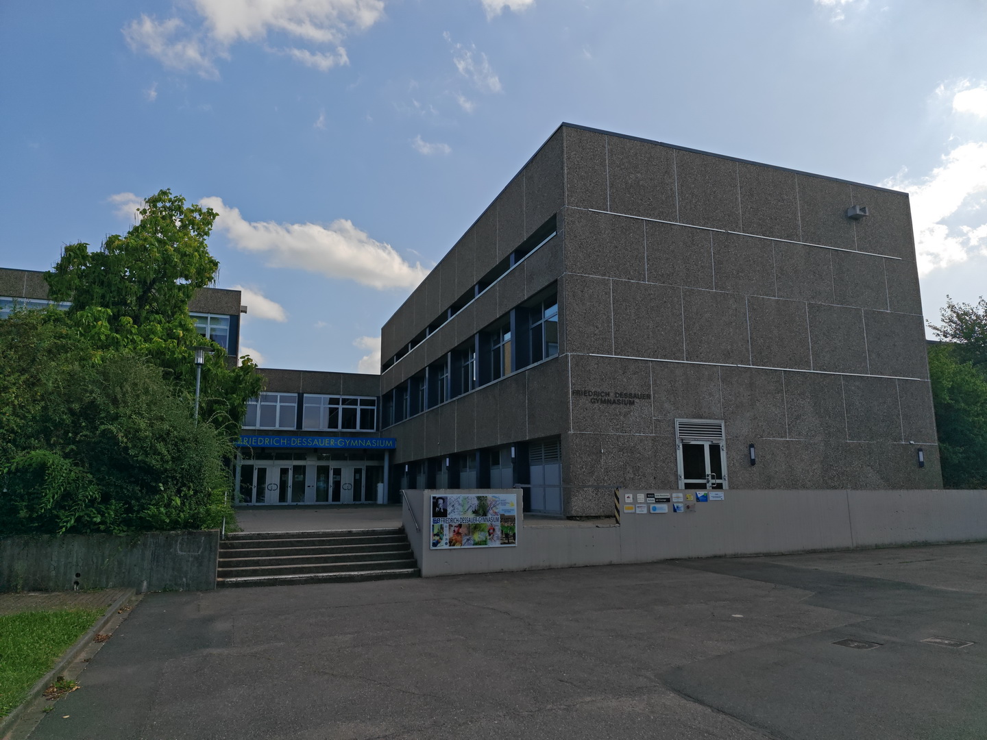 Die Außenansicht des Friedrich-Dessauer-Gymnasiums, eines markanten Gebäudes aus grauem Sichtbeton im Stil der 1970er Jahre. Eine breite Treppe führt zum Eingangsbereich des mehrstöckigen, kubischen Gebäudes. Links wird die strenge Betonarchitektur von hohen, grünen Bäumen und Sträuchern kontrastiert. Der Himmel ist blau mit weißen Wolken.
