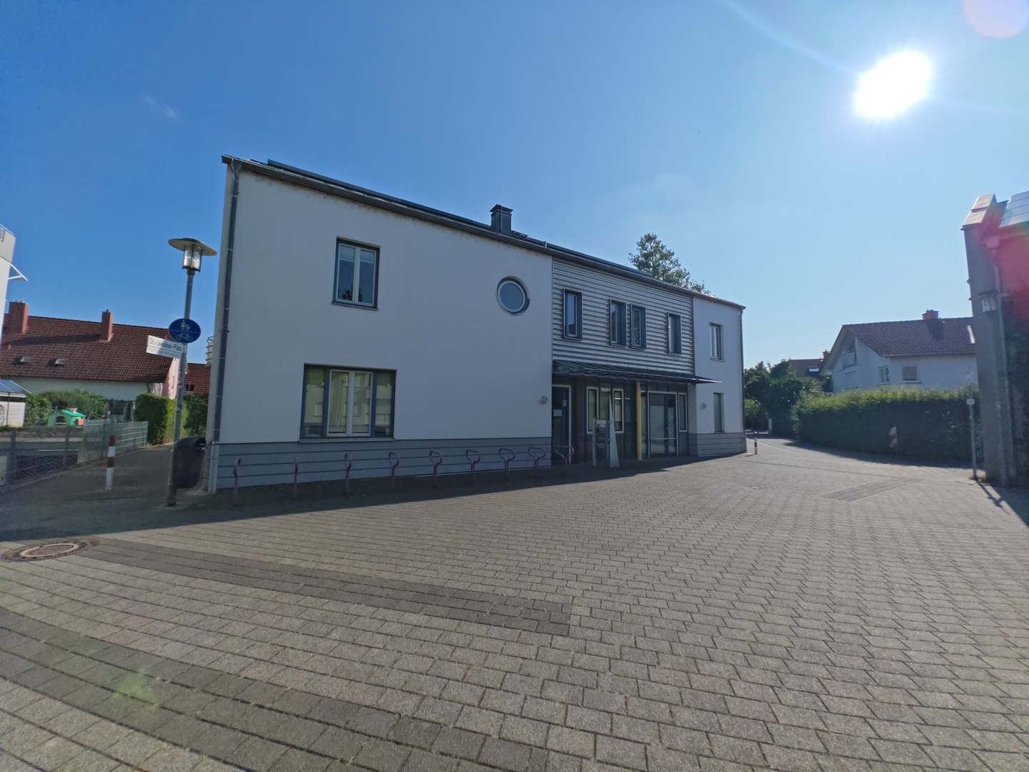 Die Außenansicht des modernen Gemeindezentrums St. Jakobus bei strahlendem Sonnenschein. Das zweistöckige Gebäude kombiniert eine weiße Putzfassade mit einer grauen Holzverkleidung im Eingangsbereich. Ein rundes Fenster im Obergeschoss ist ein markantes Detail. Davor liegt ein großer, mit grauen Steinen gepflasterter Platz.