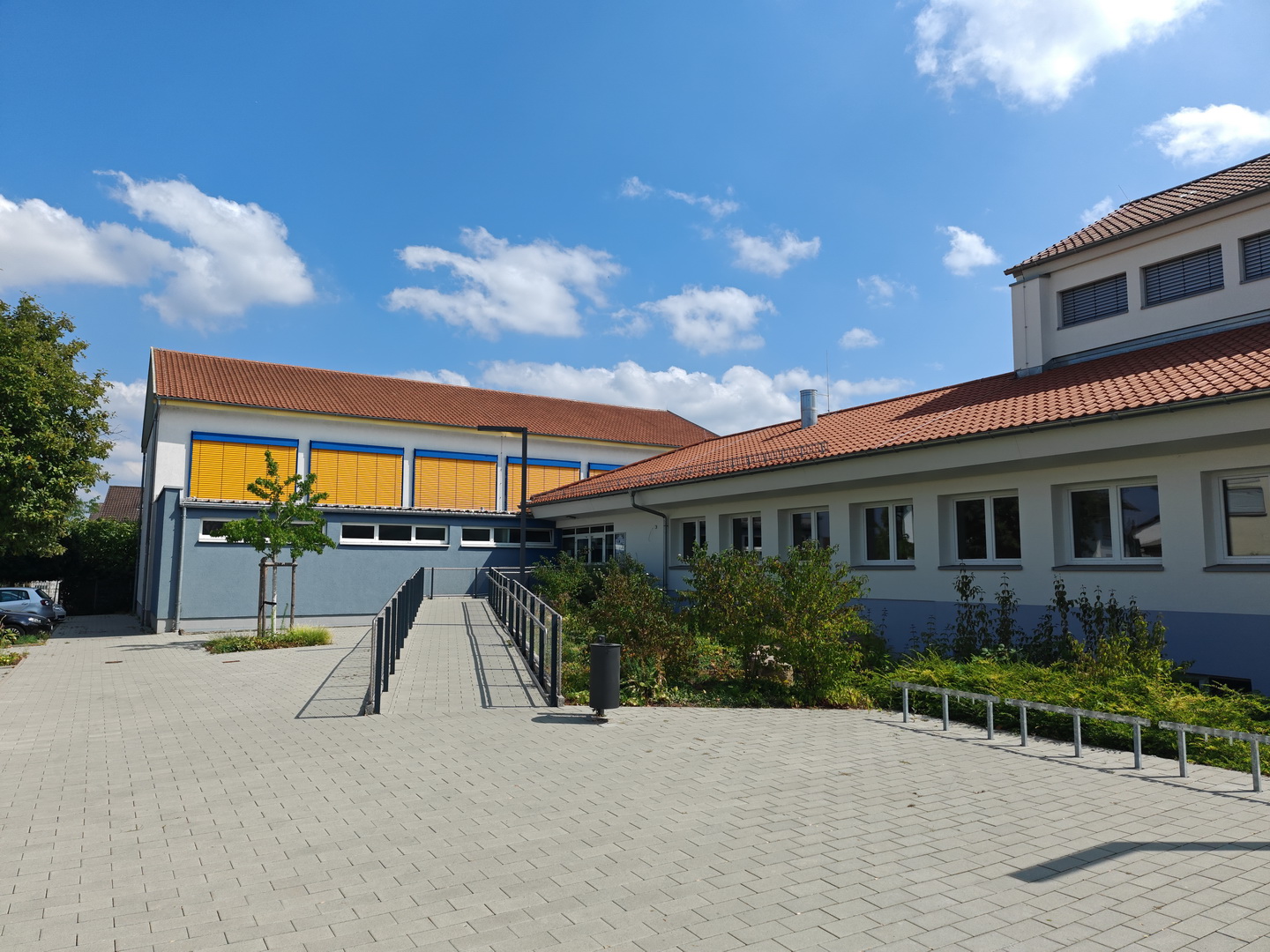 Die Außenansicht der Grundschule Großostheim unter einem blau-weißen Himmel. Eine lange, barrierefreie Rampe führt über den gepflasterten Vorplatz zum Schulgebäude. Die Fassade ist grau-blau und weiß gestrichen, und an den Fenstern eines Gebäudeteils leuchten auffällige gelbe und blaue Jalousien.