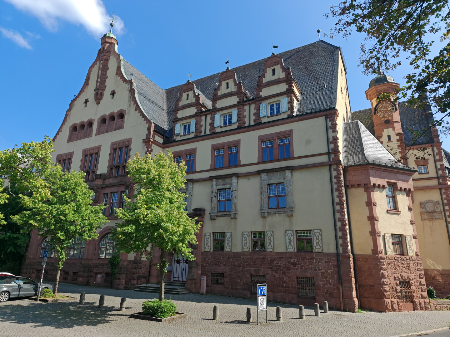 Die Außenansicht des historischen Karl-Theodor-von-Dalberg-Gymnasiums bei sonnigem Wetter. Das große, mehrstöckige Gebäude hat ein Erdgeschoss aus rotem Sandstein und eine helle obere Fassade. Die Architektur zeichnet sich durch einen markanten Stufengiebel links, ein dunkles Schieferdach mit vielen Gauben und einen kleinen Uhrturm rechts aus.