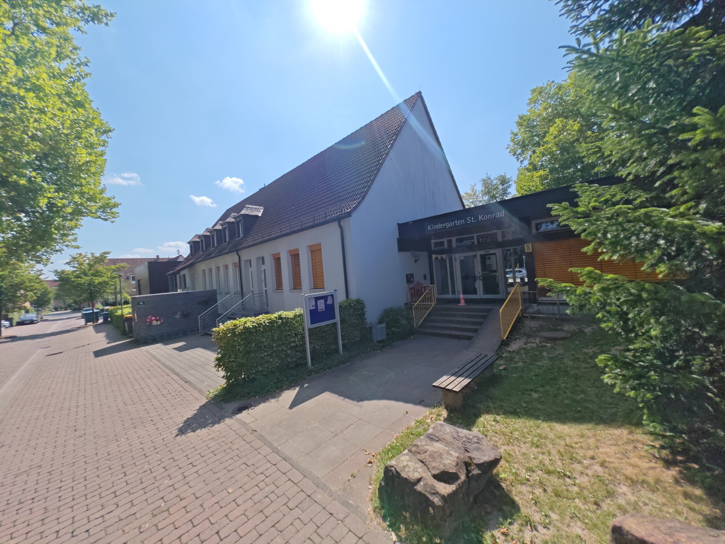 Das Gebäude des Kindergartens St. Konrad bei strahlendem Sonnenschein. Das Haus hat eine helle Fassade und ein hohes, dunkles Giebeldach. Der Eingangsbereich ist modern gestaltet mit einer Holzverkleidung und einer Treppe, die zur Glastür führt. Das Gebäude ist von grünen Bäumen und Hecken umgeben.