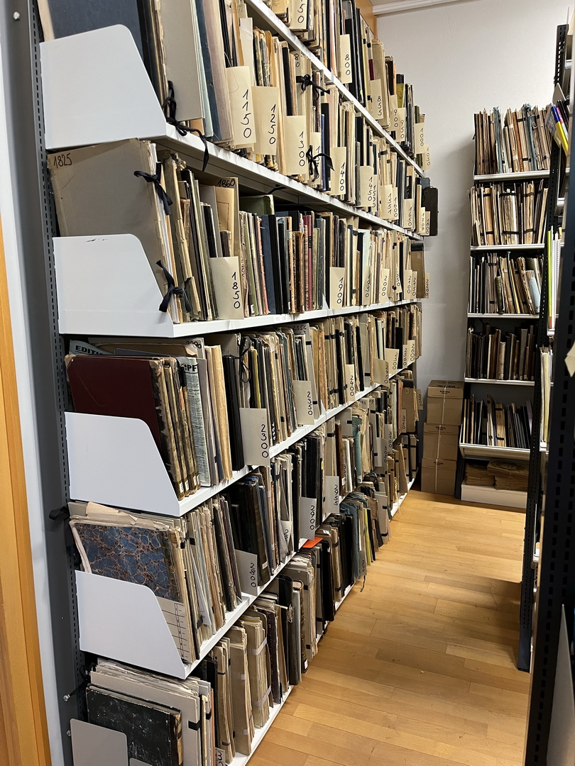Ein Blick in das Notenarchiv der Musikschule. Lange Regalreihen sind bis unter die Decke mit alten, gebundenen Notenbüchern gefüllt. An vielen Bänden stecken Zettel mit historischen Jahreszahlen.