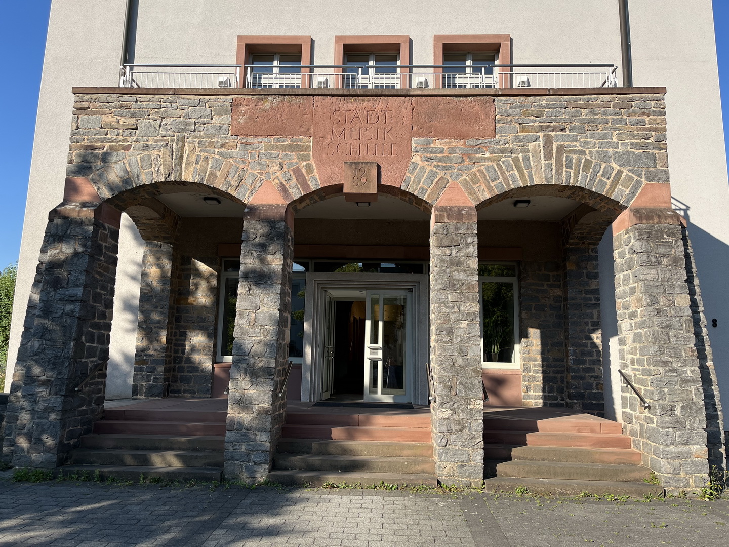 Eine Nahaufnahme des Eingangs der Städtischen Musikschule Aschaffenburg. Der Eingang besteht aus drei Bögen, die von wuchtigen Steinsäulen getragen werden. Über dem mittleren Bogen ist "STÄDT. MUSIKSCHULE" in Stein gemeißelt.