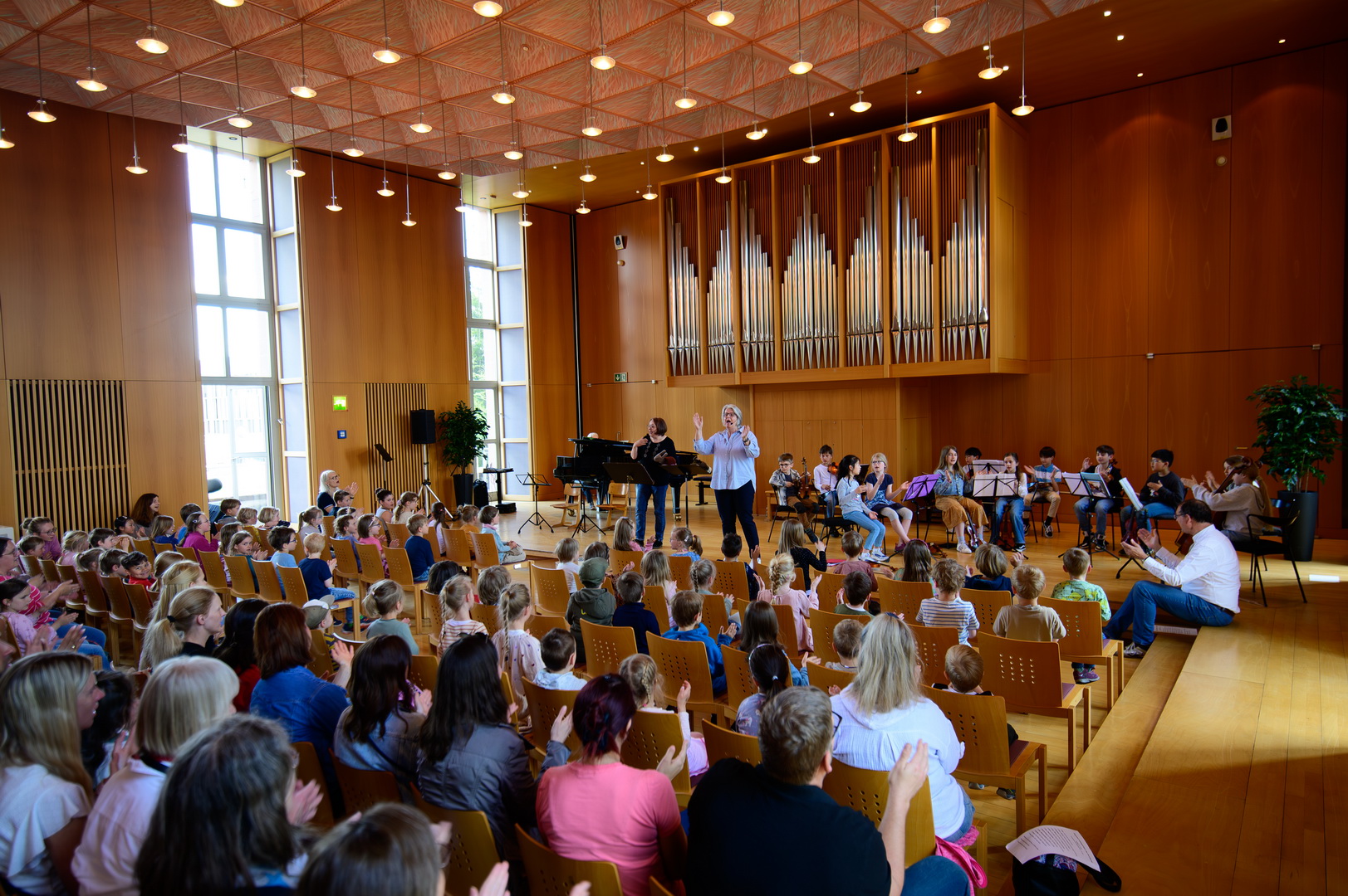 Ein großer Konzertsaal mit Holzverkleidung, gefüllt mit einem Publikum aus Kindern und Erwachsenen. Auf der Bühne musiziert eine Gruppe von Kindern und Erwachsenen. Im Hintergrund der Bühne steht eine große Orgel.