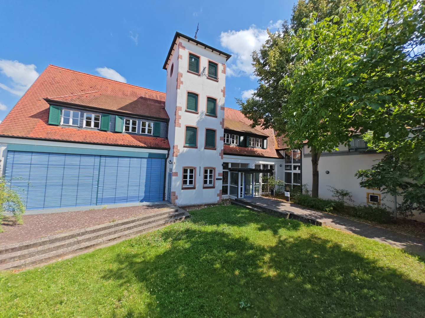 Die Außenansicht der Musikschule Großostheim an einem sonnigen Tag. Das Gebäude hat eine helle Fassade, ein markantes rotes Ziegeldach und einen zentralen Turm. Viele der Fenster sind mit grünen Klappläden versehen. Im Vordergrund befindet sich eine kleine Rasenfläche mit Stufen, die zum Gebäude führen.