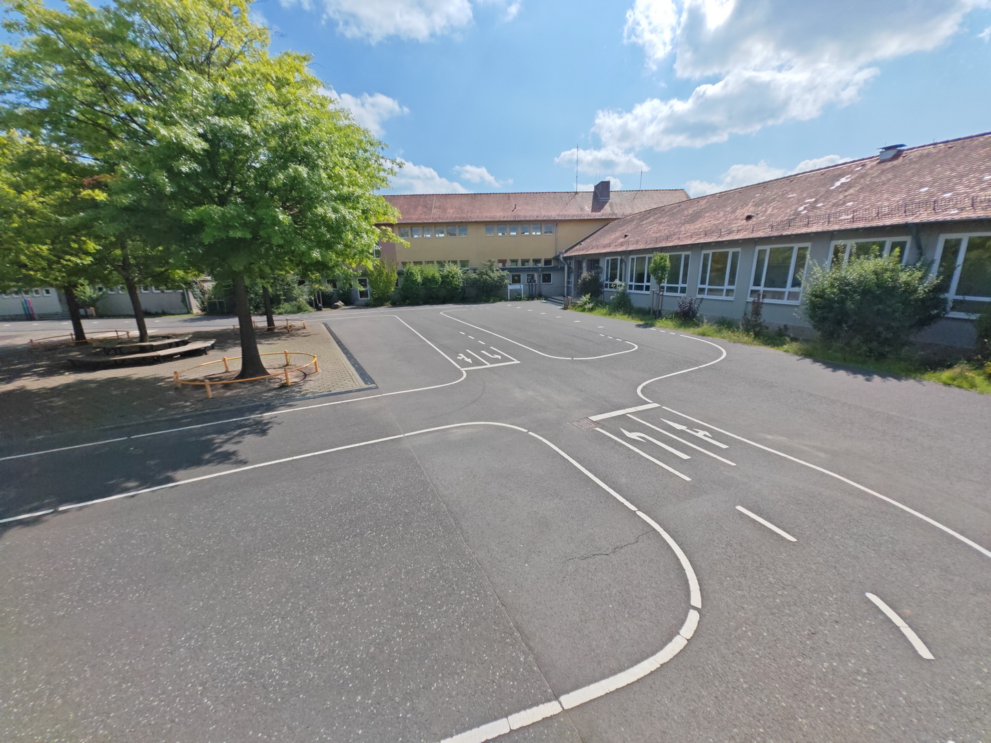 Der Schulhof der Pestalozzi-Grundschule, der als Verkehrsübungsplatz gestaltet ist. Auf dem asphaltierten Boden im Vordergrund sind weiße Fahrbahnmarkierungen aufgemalt. Im Hintergrund steht das Schulgebäude unter einem sonnigen, leicht bewölkten Himmel. Am linken Bildrand sind grüne Laubbäume zu sehen.