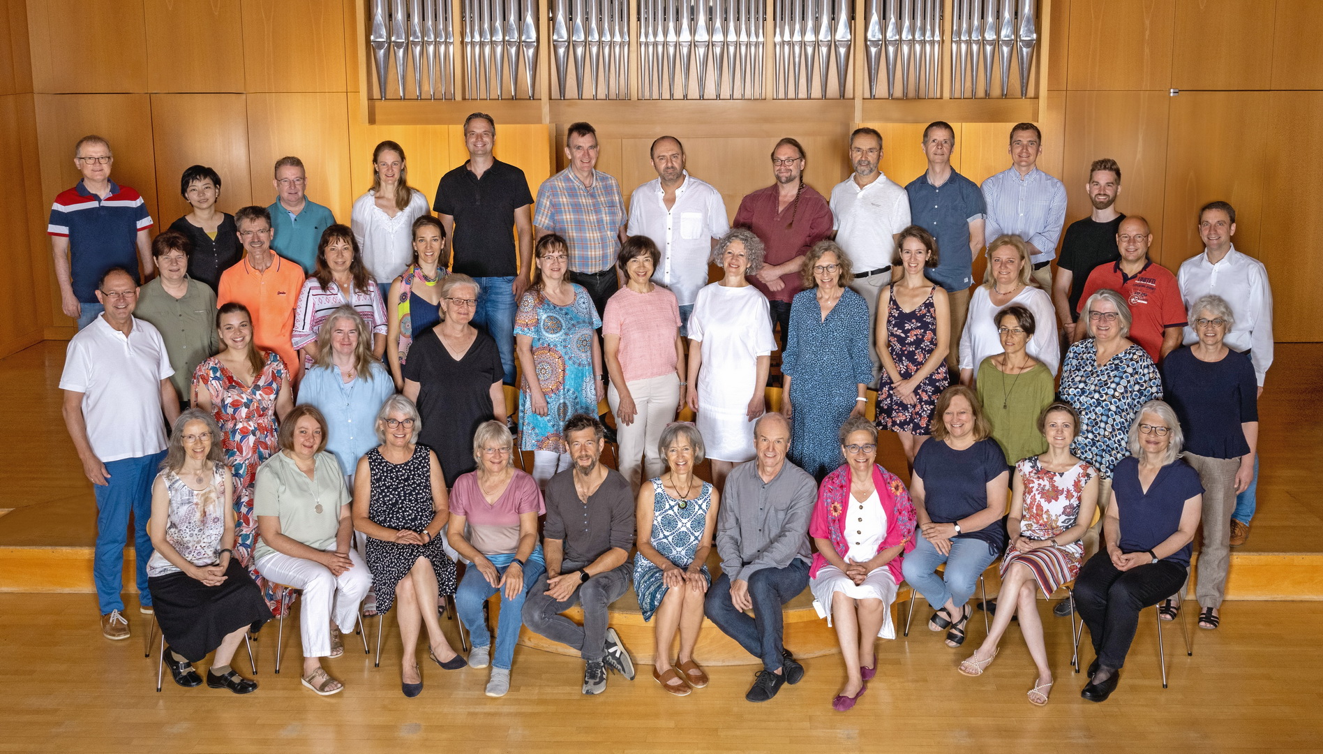 Ein Gruppenfoto des Kollegiums der Städtischen Musikschule Aschaffenburg. Eine große Gruppe erwachsener Männer und Frauen posiert in vier Reihen in einem Konzertsaal. Im Hintergrund ist die große Pfeifenfront einer Orgel zu sehen.