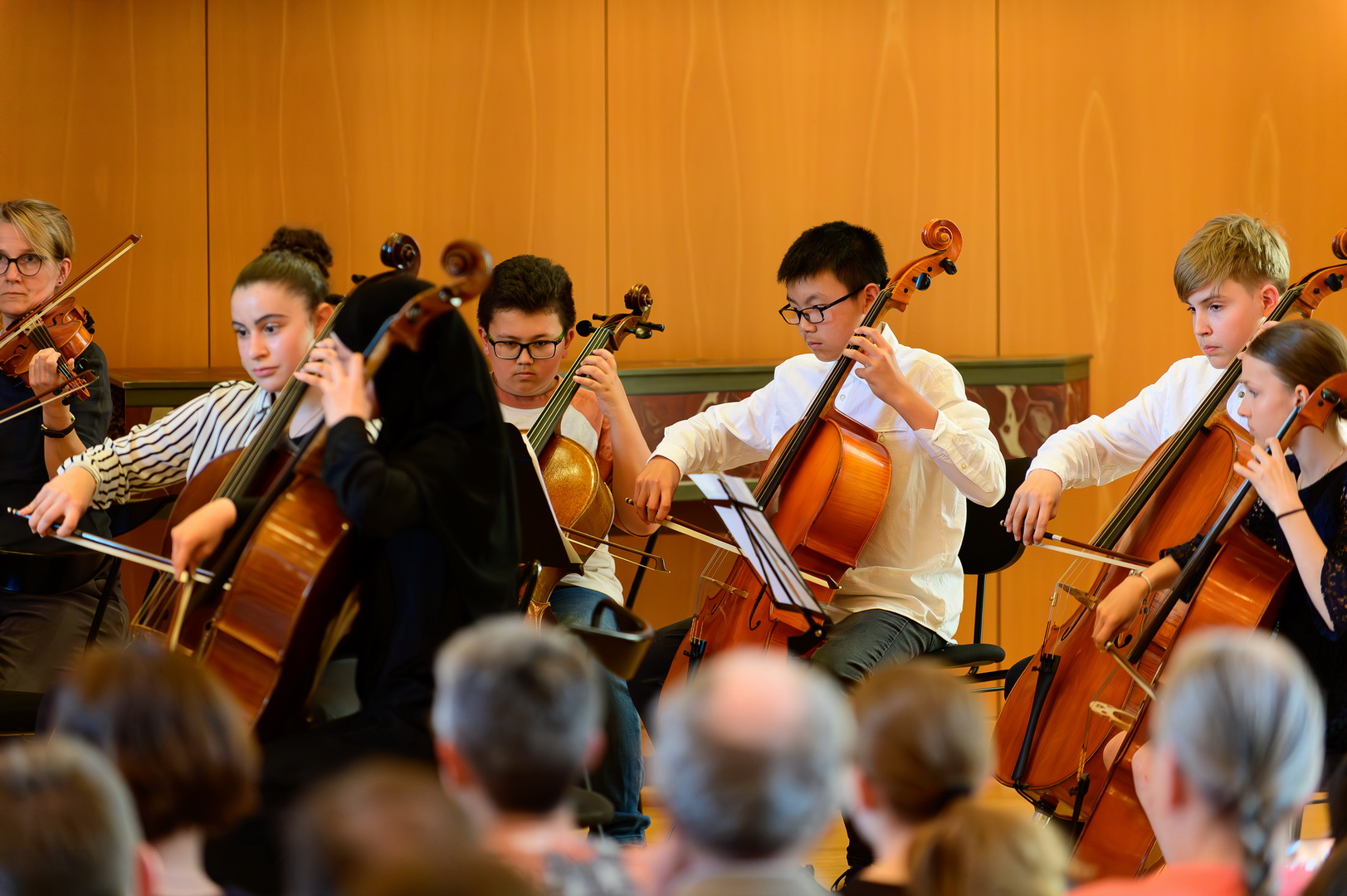 Ein Streicher-Ensemble aus jungen Musizierenden bei einem Auftritt. Mehrere Cellisten und eine Geigerin spielen konzentriert auf ihre Instrumente. Im unscharfen Vordergrund sind die Köpfe des Publikums zu sehen.