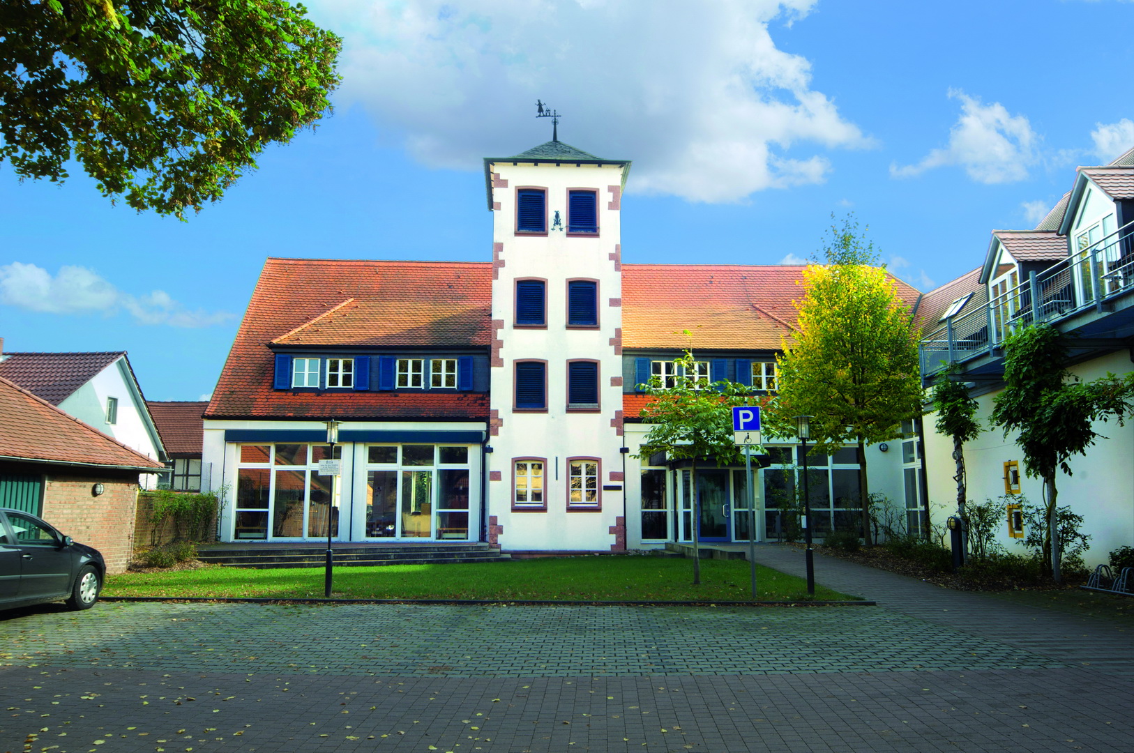 Die Frontansicht der Städtischen Musikschule Aschaffenburg an einem sonnigen Tag. Das Gebäude hat einen markanten, weißen Turm in der Mitte, ein rotes Ziegeldach und blaue Fensterläden. Davor befindet sich ein gepflasterter Parkplatz und eine kleine Rasenfläche.