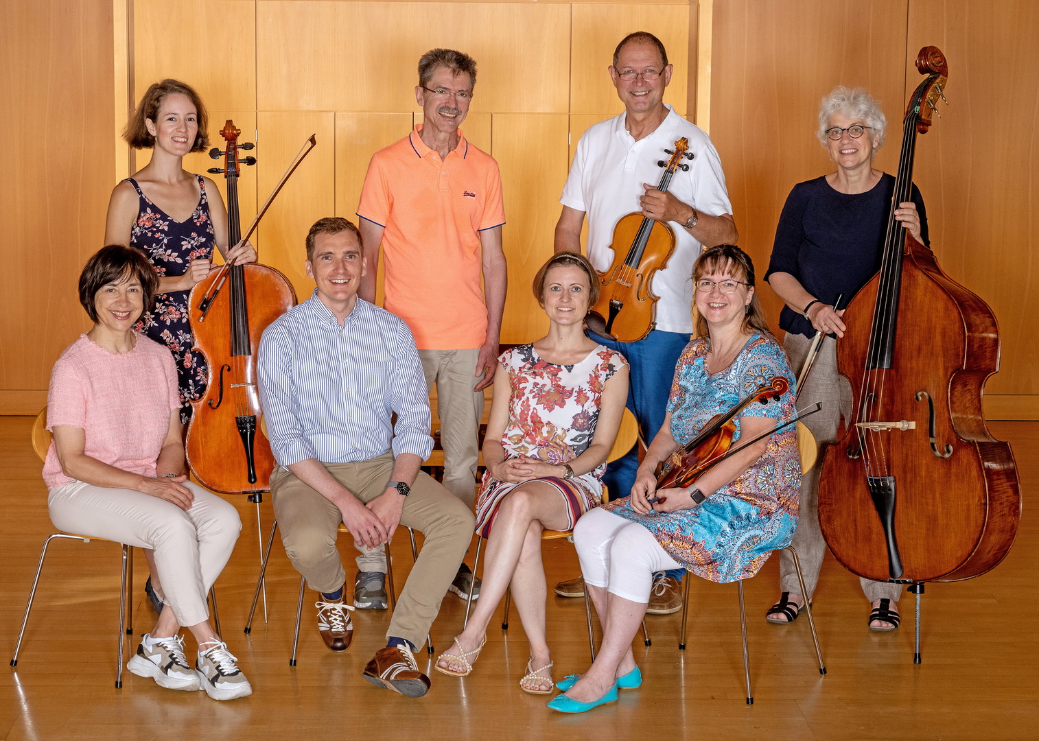 Ein Gruppenfoto der achtköpfigen Fachschaft für Streichinstrumente. Die Lehrkräfte, Männer und Frauen, posieren teils sitzend, teils stehend in einem holzvertäfelten Saal. Sie präsentieren ihre Instrumente: Geigen, ein Cello und ein Kontrabass.