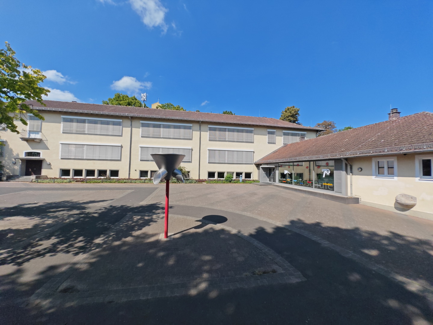 Außenansicht der Strietwald-Schule an einem sonnigen Tag. Der Blick geht über den gepflasterten Schulhof auf das mehrstöckige, helle Schulgebäude im Hintergrund. Im Vordergrund des Hofes steht eine moderne Skulptur mit einem roten Standfuß. Rechts ist ein flacherer Anbau mit einer Glasfront zu sehen. Der Himmel ist blau mit einigen wenigen Wolken.