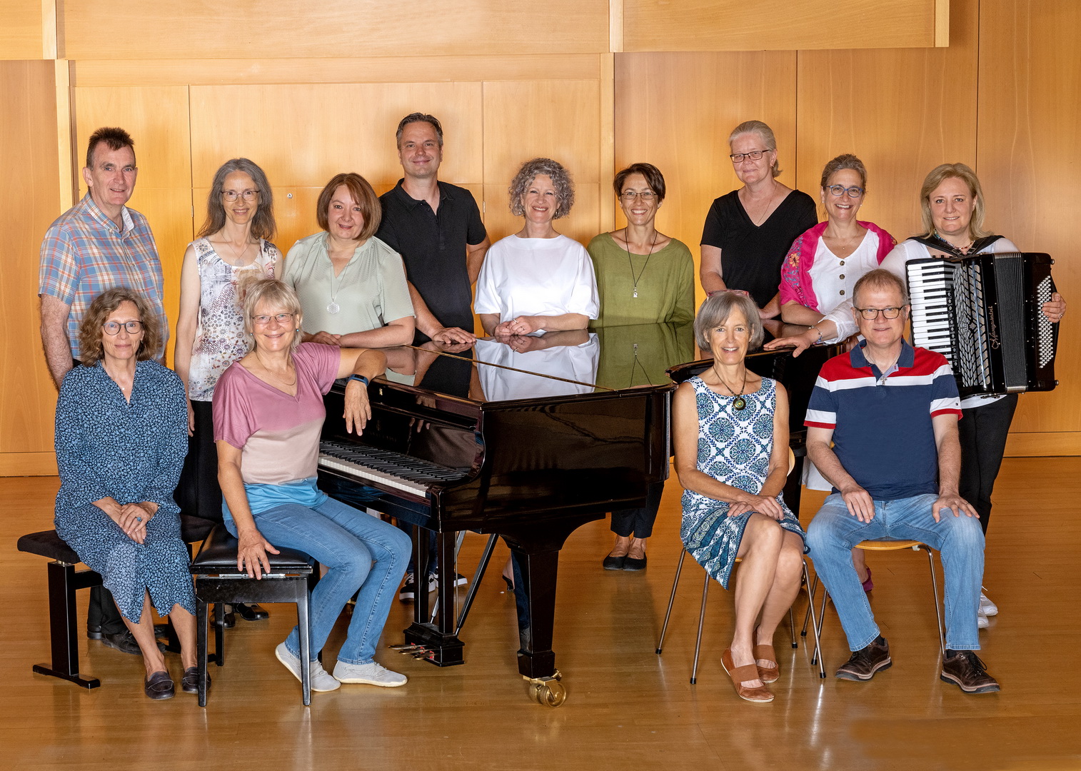 Ein Gruppenfoto der Lehrkräfte für Tasteninstrumente. Eine Gruppe von Männern und Frauen ist in einem holzvertäfelten Saal um einen schwarzen Flügel herum arrangiert. Eine Lehrerin am rechten Rand hält ein Akkordeon.