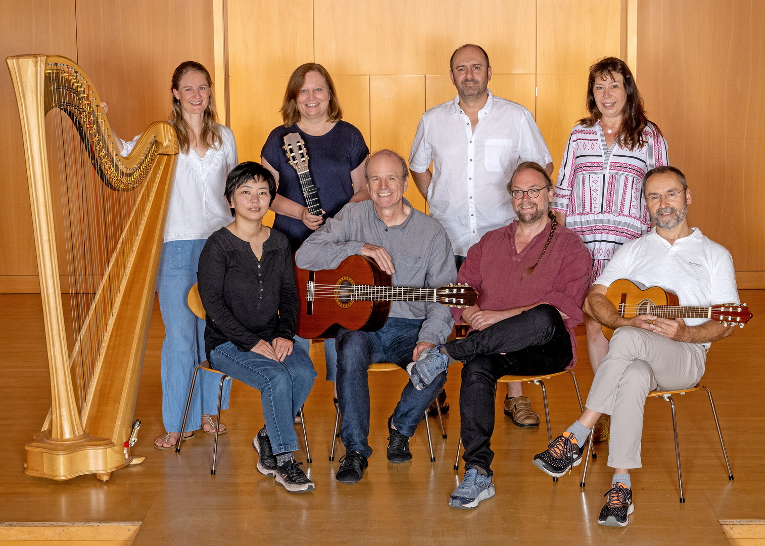Ein Gruppenfoto der achtköpfigen Fachschaft für Zupfinstrumente. Vier Frauen und vier Männer posieren teils sitzend, teils stehend mit ihren Instrumenten – einer Harfe und mehreren Gitarren – in einem holzvertäfelten Saal.